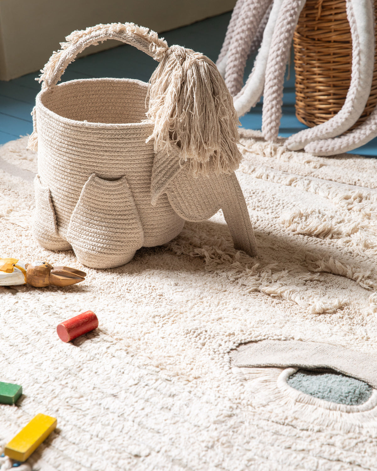 Panier Mammouth posé sur un tapis crème en relief au milieu de cubes en bois, avec un panier tressé en arrière-plan