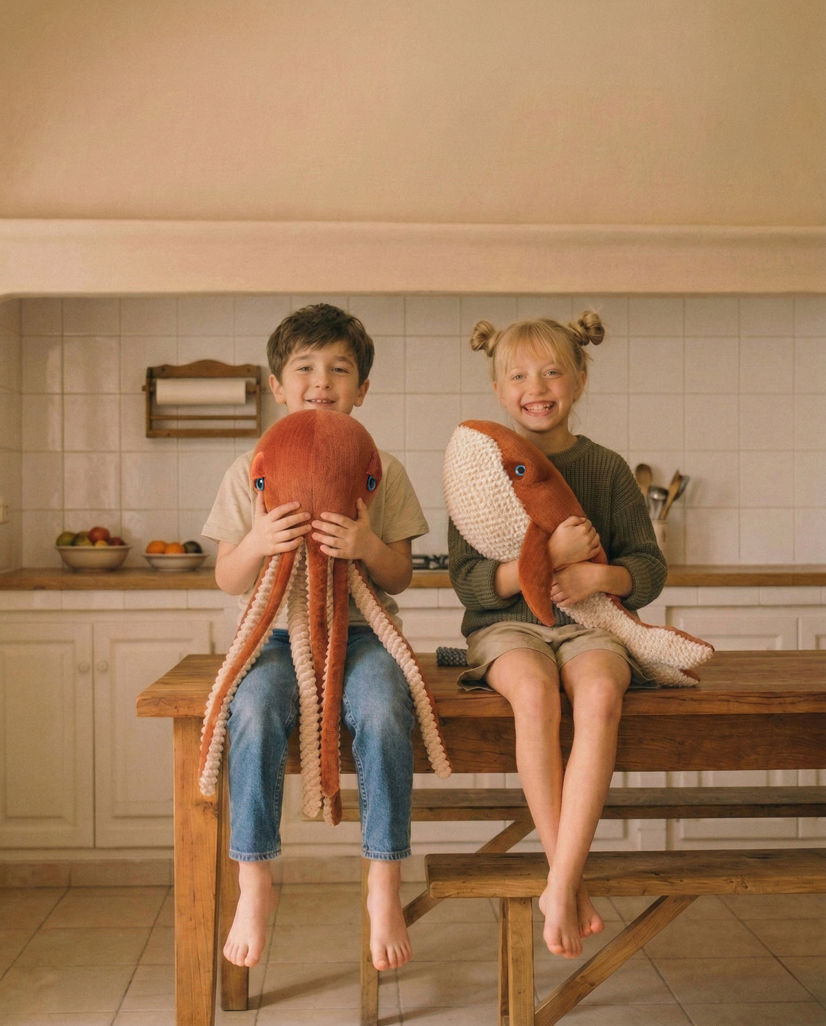 Deux enfants souriants assis sur une table de cuisine, serrant une pieuvre rouge et une baleine rouge en peluche